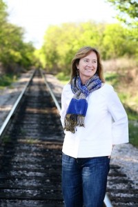 Cynthia dressed in jeans and a white blouse and scarf standing on railroad tracks